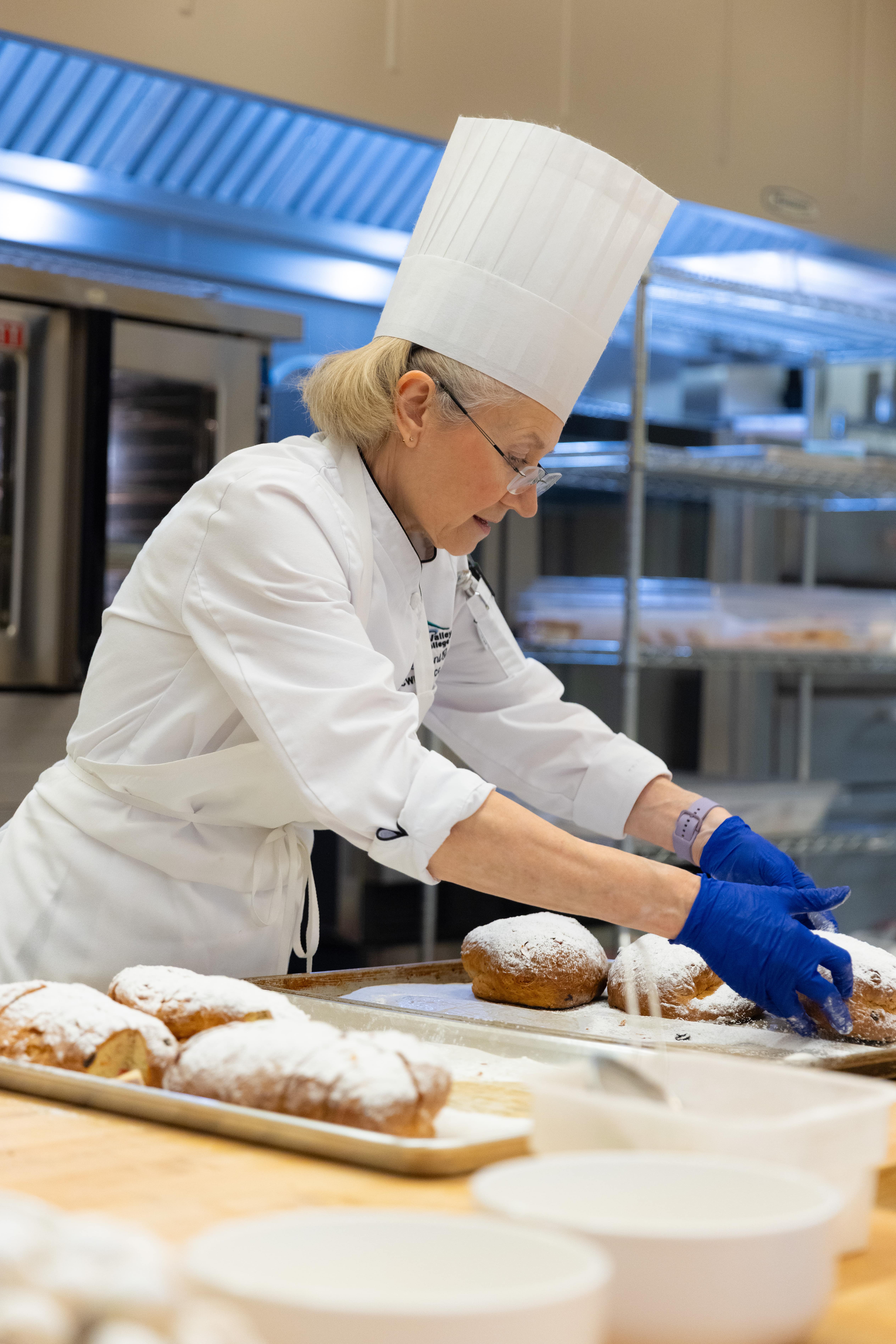 A group of six chefs, some in uniforms and others in casual attire, stand behind a table laden with various breads and dishes. They are smiling warmly.
